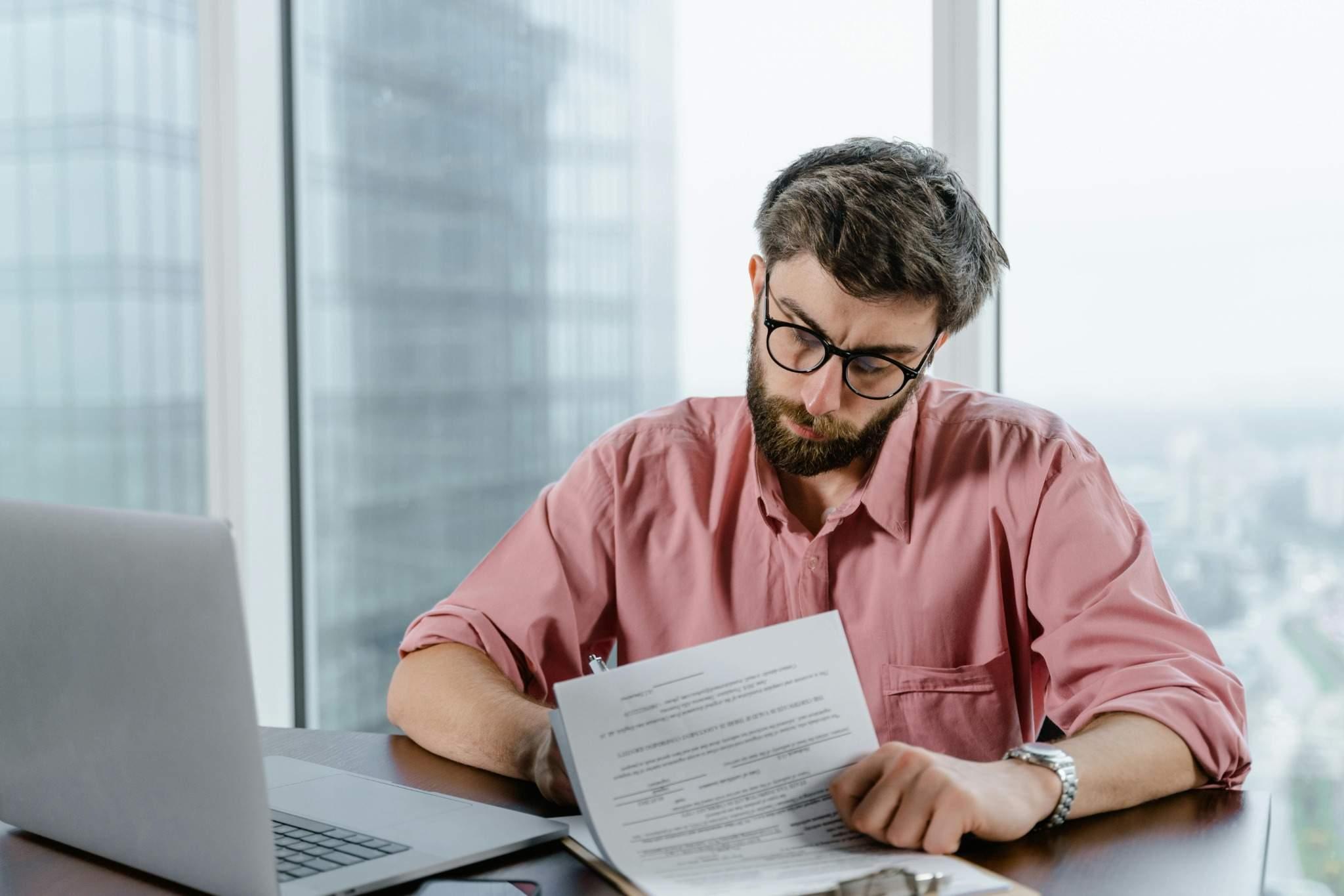 hombre con gafas y una camisa rosada consultando unos documentos en papel con un ordenador encendido enfrente.