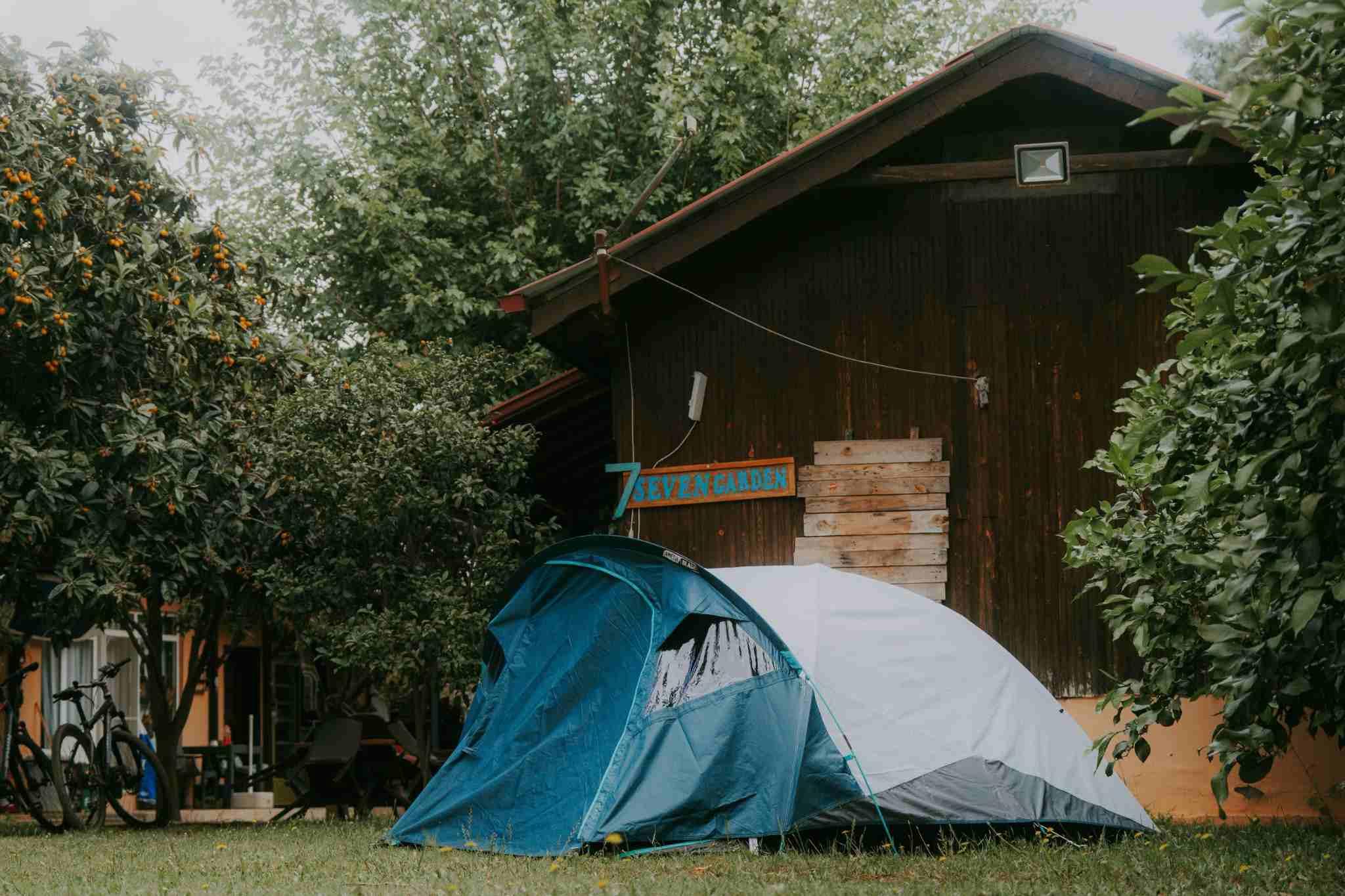 tienda de campa&ntilde;a azul situada junto a un bungalow de madera en un camping rodeado de vegetaci&oacute;n.