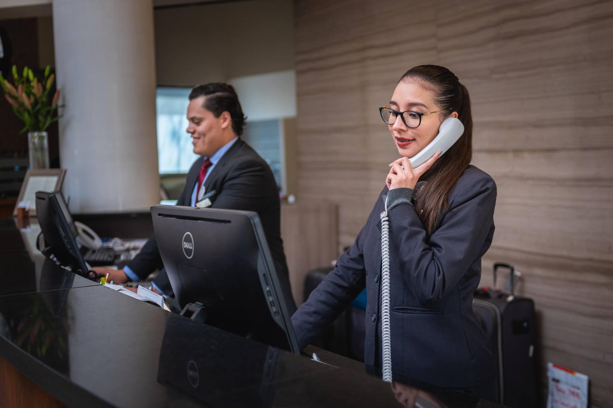 dos trabajadores de un hotel en la recepci&oacute;n, una de ellas hablando por tel&eacute;fono y el otro consultando el ordenador.