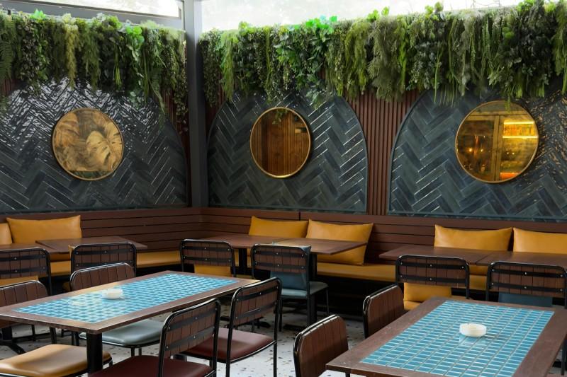 terraza interior de un restaurante con varias mesas rectangulares y sillas con respaldo, con vegetaci&oacute;n y decoraci&oacute;n moderna.